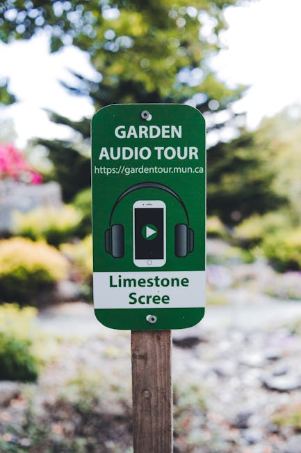 A close-up of a green garden sign mounted on a wooden stake, indicating an audio garden tour with the website link 'https://gardentour.mun.ca'. The sign features an illustration of headphones and a smartphone showing a play button, with text that reads 'GARDEN AUDIO TOUR' and 'Limestone Screen'. In the background, blurred foliage, trees with lush green leaves, and sunlight filtering through the canopy are visible, suggesting a well-maintained garden space typical of outdoor landscaping near Belsize Park. The garden area appears tidy with varied plant textures, including leafy bushes and grassy sections, possibly part of a landscape maintenance or garden care project by Gardeners Belsize Park.